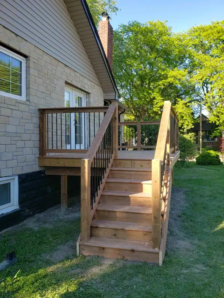 Wooden deck with railing and stairs leading down to a grassy backyard, attached to a stone and siding house with French doors.