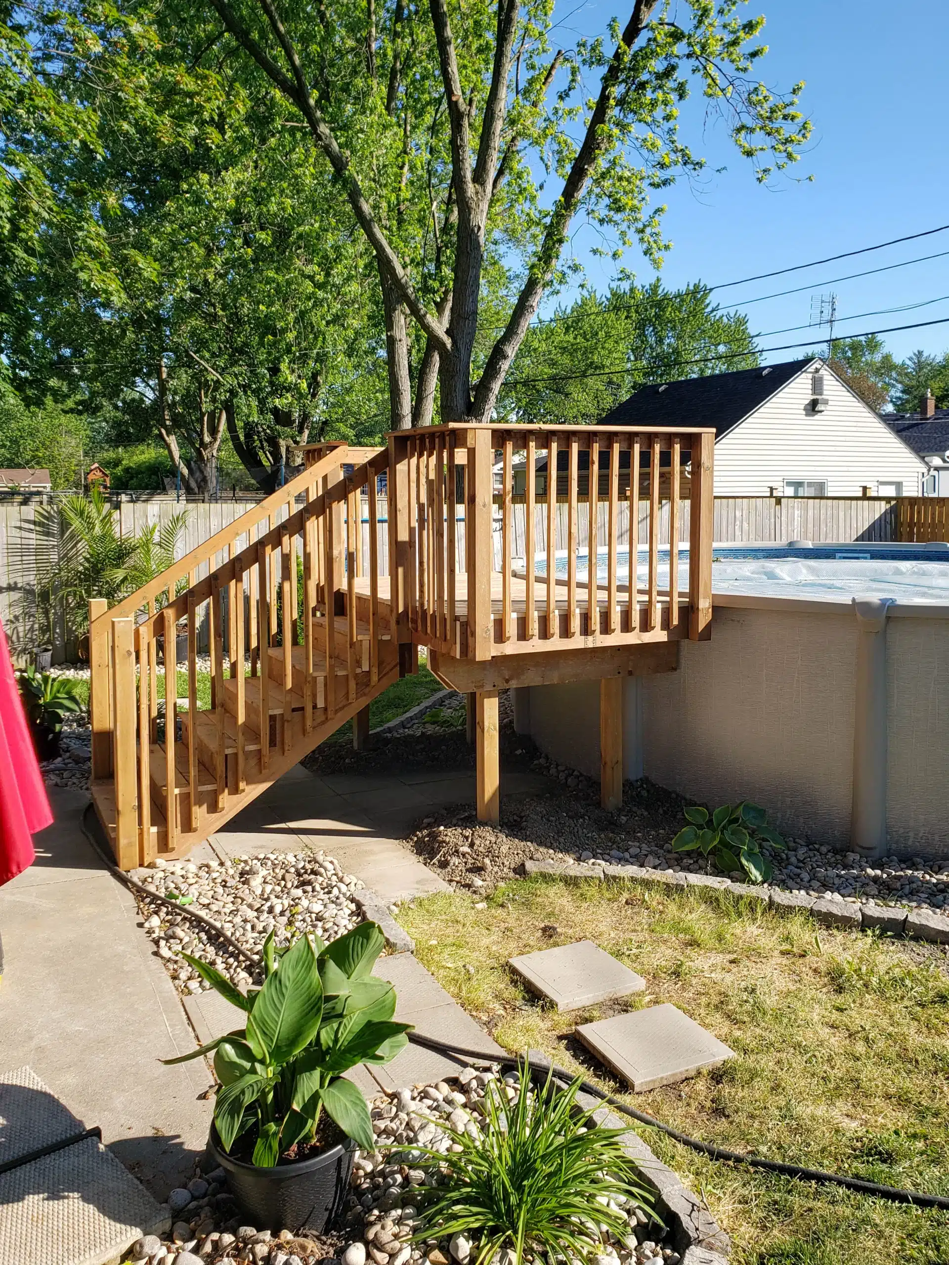 Above-ground swimming pool with attached wooden deck and stairs, surrounded by a landscaped backyard with stone walkway, plants, and trees.