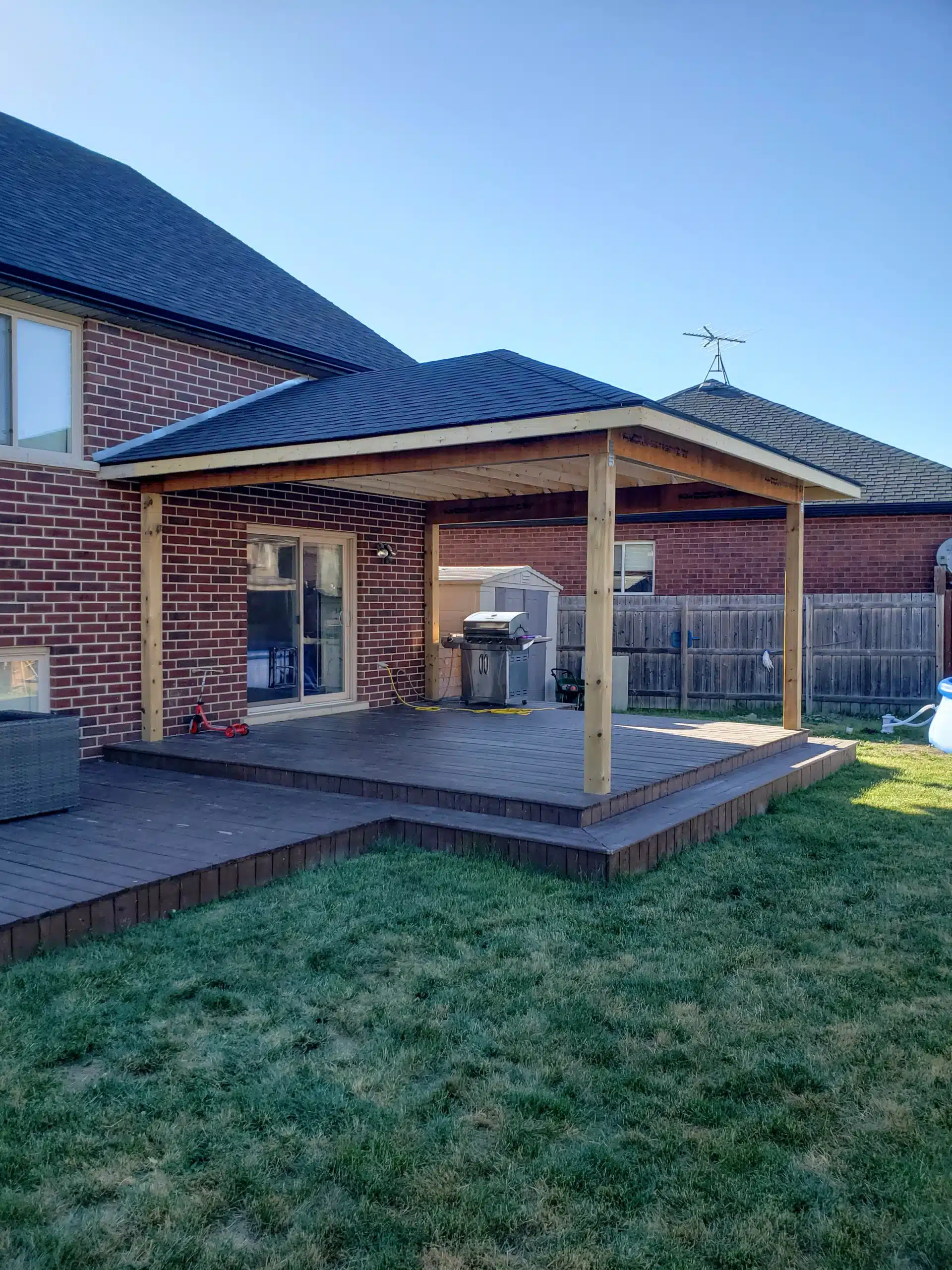Backyard wooden deck with a covered roof structure attached to a brick house, featuring open grassy lawn and patio furniture.