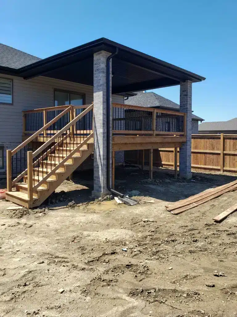Covered wooden deck with railing and staircase attached to a brick and siding house, built above ground level in a fenced backyard under construction.