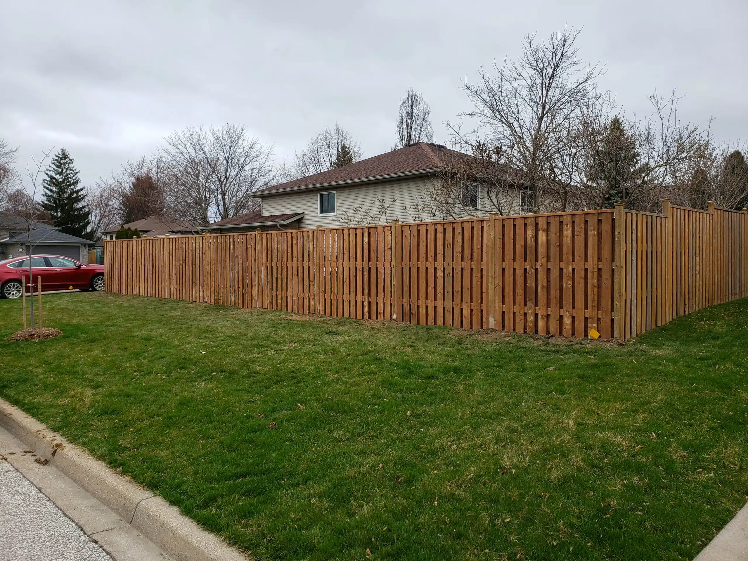 Wooden privacy fence with vertical panels enclosing a suburban backyard, with a grassy lawn, red car, and neighboring houses in the background.