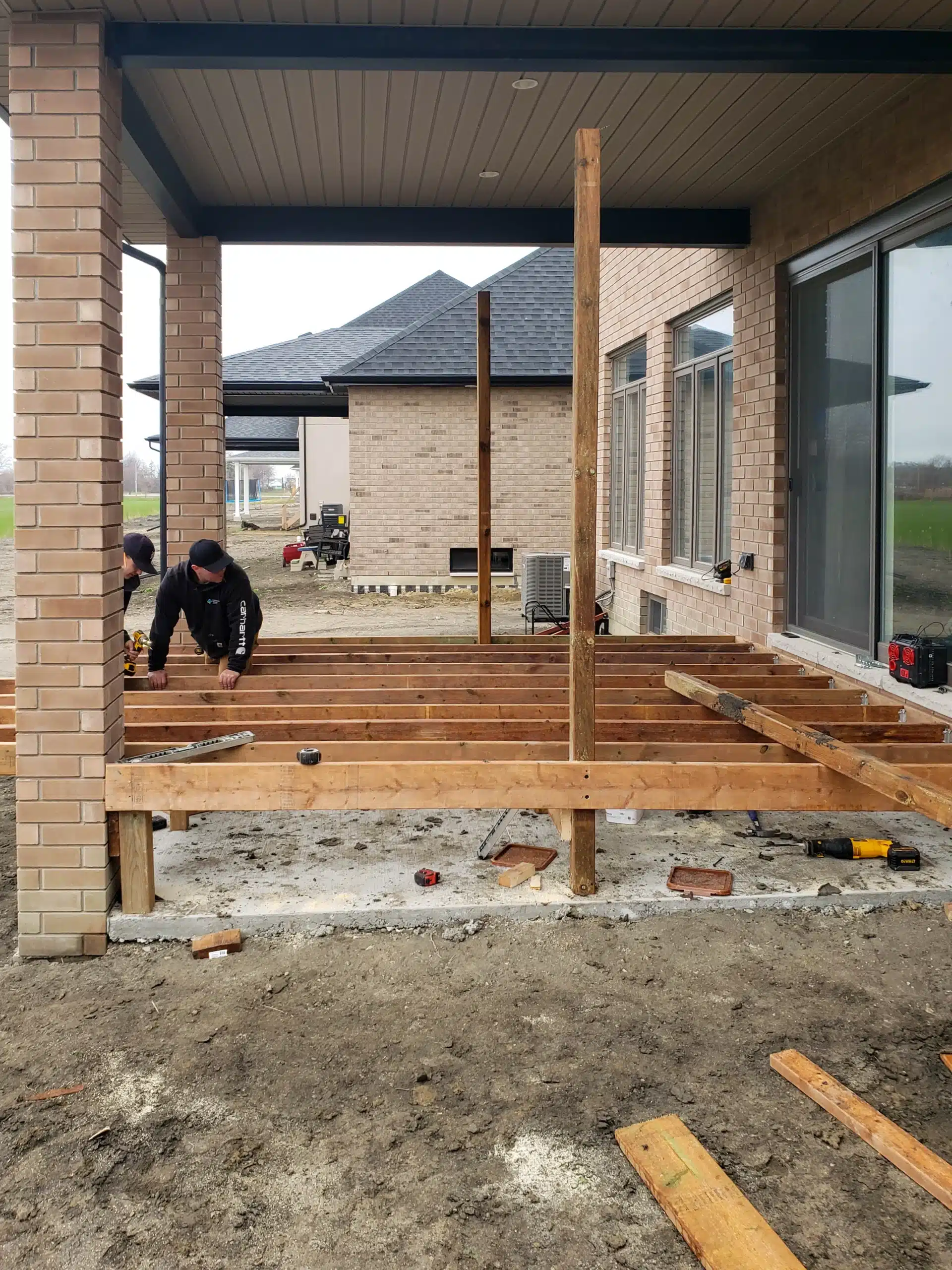 Construction of a wooden deck in progress at the back of a brick house, with workers installing support beams under a covered patio area.